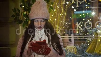 美丽迷人的女孩站在一家装饰有花环的橱窗附近，使用智能手机。 新年圣诞节